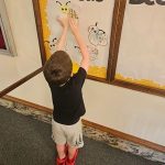Pre-school aged boy hangs up a paper bee he colored in a display case to show his participation in the library's 1000 Books Before Kindergarten reading challenge