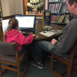 A little girl sits at one of the computer stations in the childrens library with her dad watching her use it