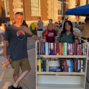 Man leans on book cart while woman fans out cash they won at the library's Night market table outside the Library
