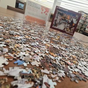 puzzle pieces are scattered around the top of a table with the puzzle box and sign indicating that this is a puzzle that all library visitors can put together