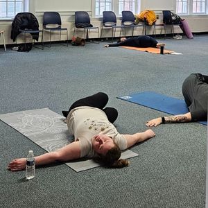 a Yoga instructor is seen in the distance while 2 women practice the pose she is instructing in the community room at the library
