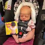 2 week old baby is strapped into a car seat with a board book placed beside her