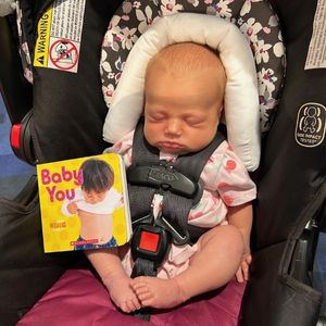 2 week old baby is strapped into a car seat with a board book placed beside her